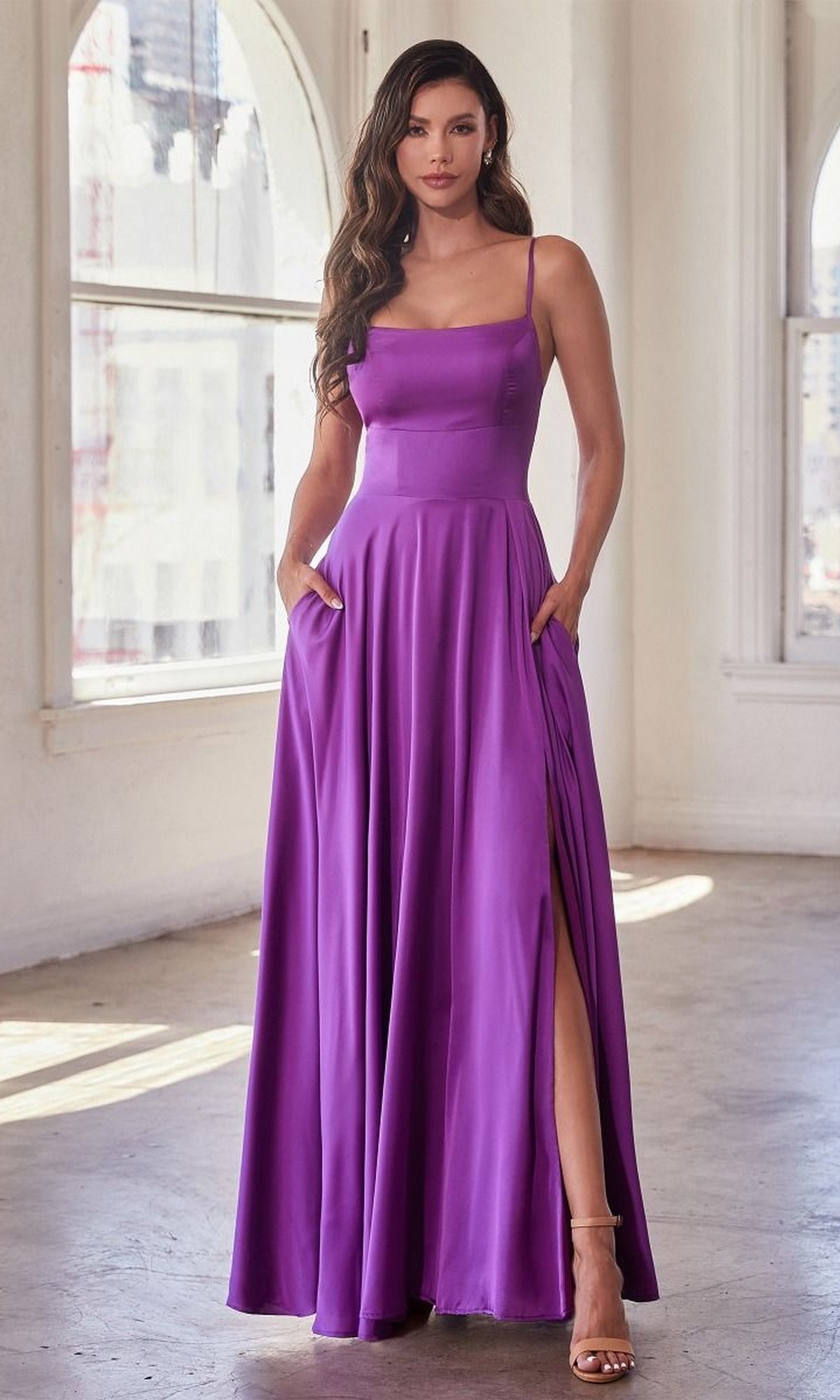 A woman stands indoors wearing the Lace-Up Classic Long Prom Dress B8402 in purple, featuring thin straps, a high slit, and side pockets; she poses confidently with one leg slightly forward.